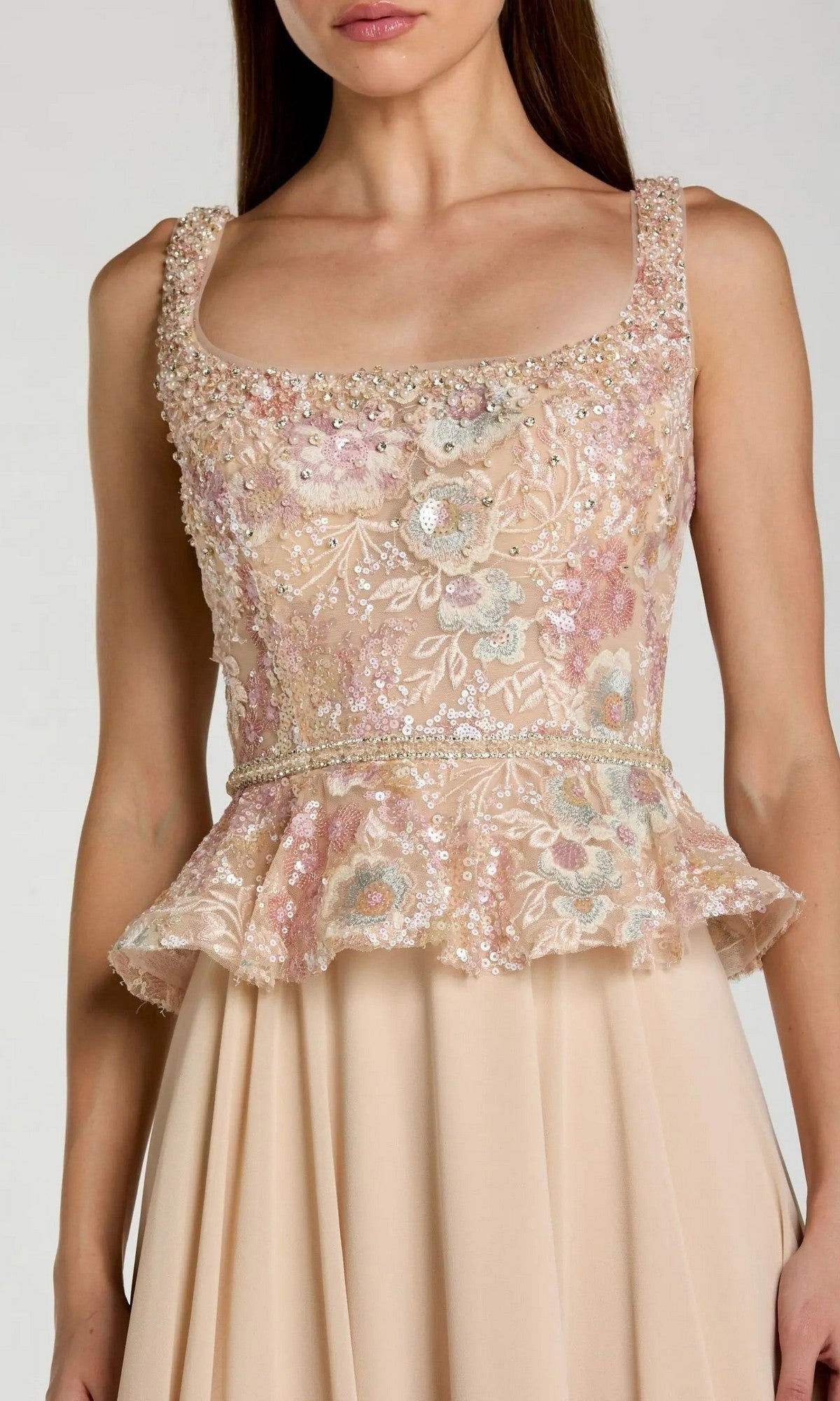 A woman wearing the Mac Duggal 21317 long formal dress, which features a sleeveless, light pink design with a floral embroidered bodice, delicate pearl embellishments, and a flattering peplum waistline.